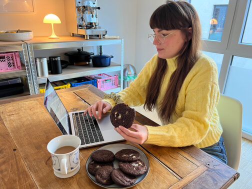 Vanessa sitzt am Küchentisch mit schwarzen Tee und Cookies und tippt in ihrem Laptop.